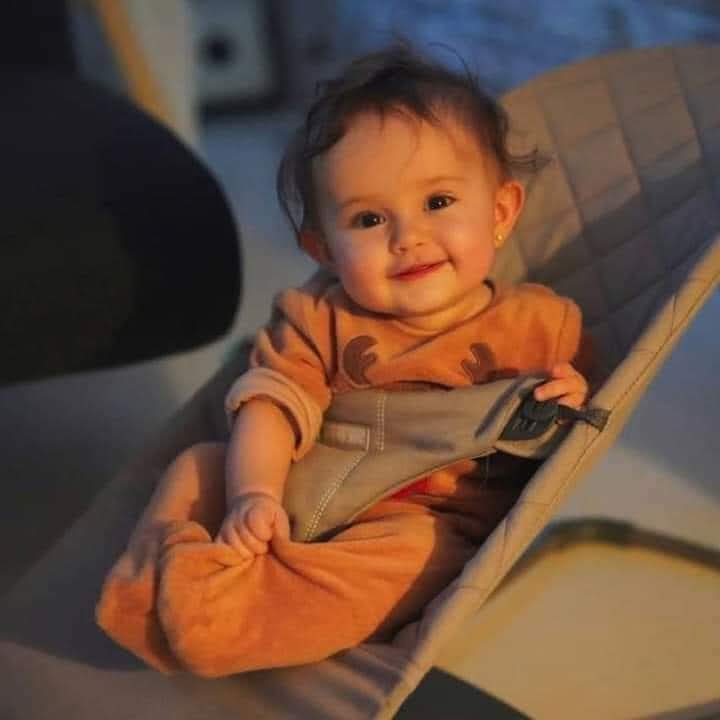 Cheerful smiling baby in an orange onesie seated in a padded grey baby bouncer under warm soft in...