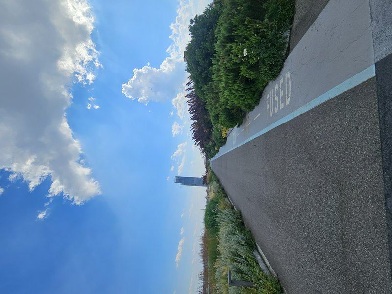 Scenic paved path with blue line marking, green bushes on one side, under a bright blue sky with white clouds.