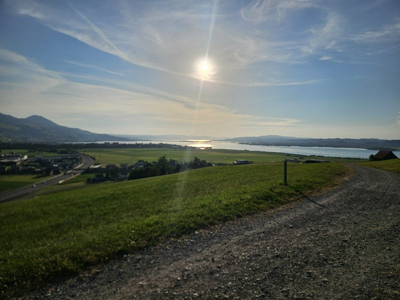 Elevated hillside gravel path overlooking a green rural valley, calm lake, and distant mountains under bright sunlit sky