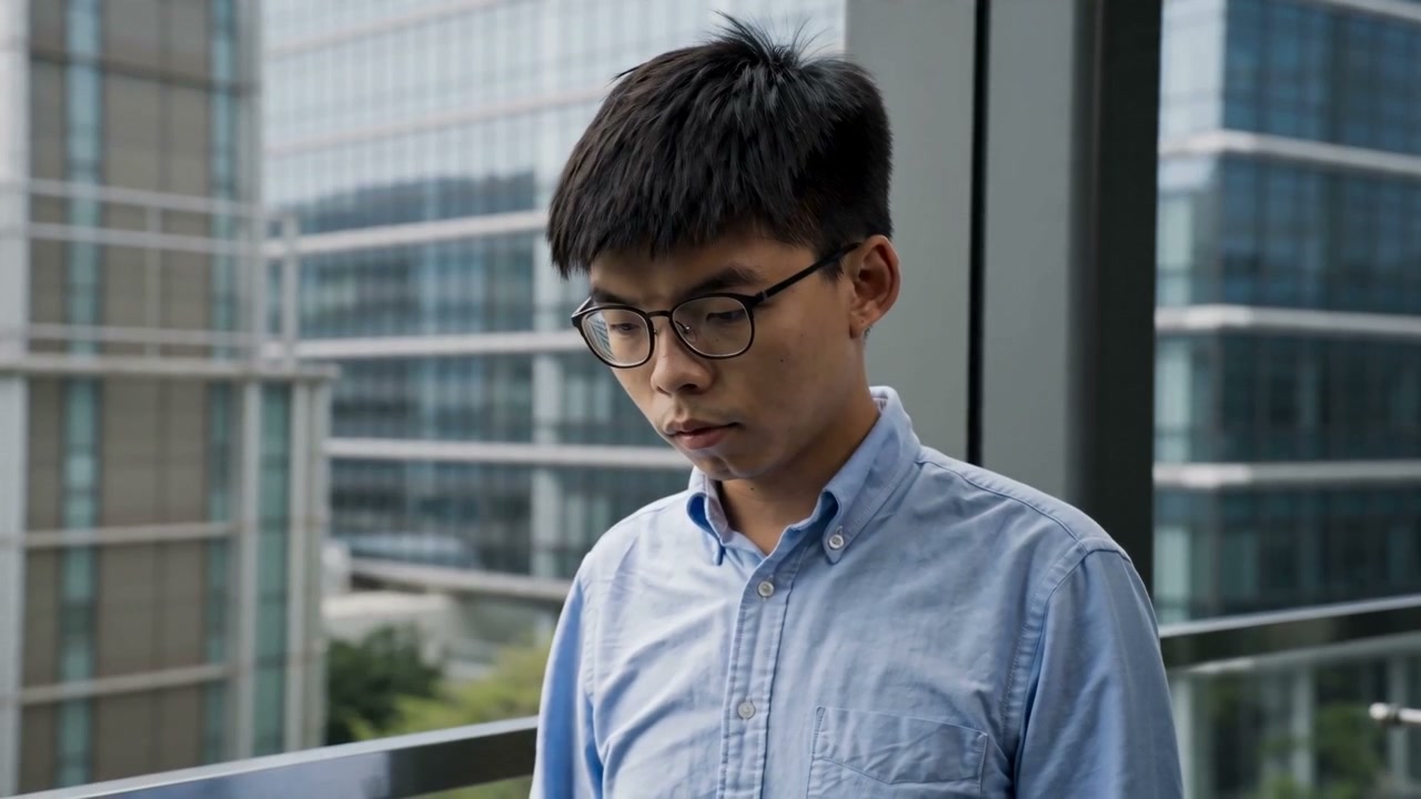 Asian man with glasses in a light blue shirt standing on a balcony overlooking modern skyscrapers.
