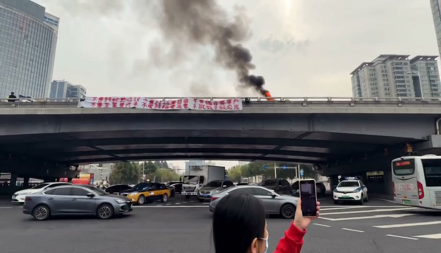 A person records a car fire on an overpass as smoke and flames rise above a busy city street.