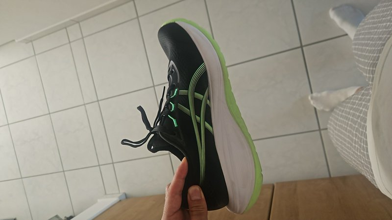 Black athletic shoe with neon green sole and laces being held indoors over tiled floor.