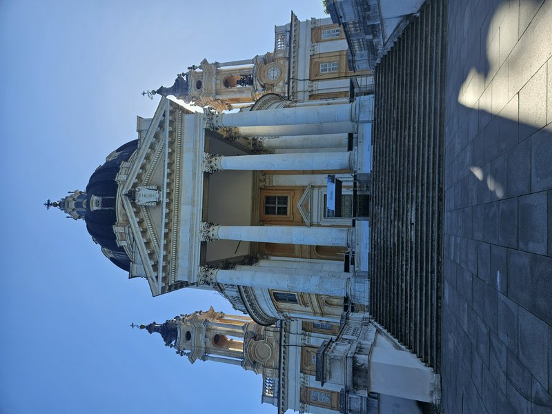 Grand neoclassical building with tall columns, ornate domes, and stone stairs under a blue sky.