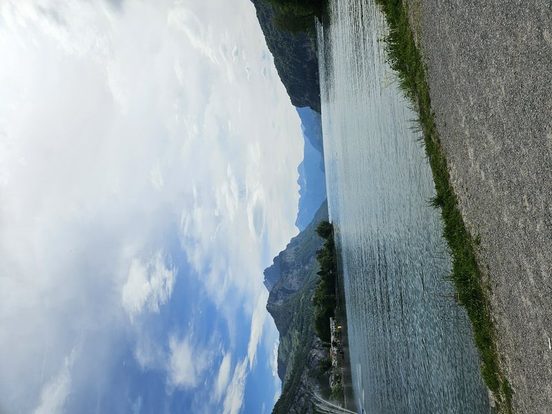 Wide mountain lake with blue water, pebbled shoreline, and forested cliffs under a cloudy sky