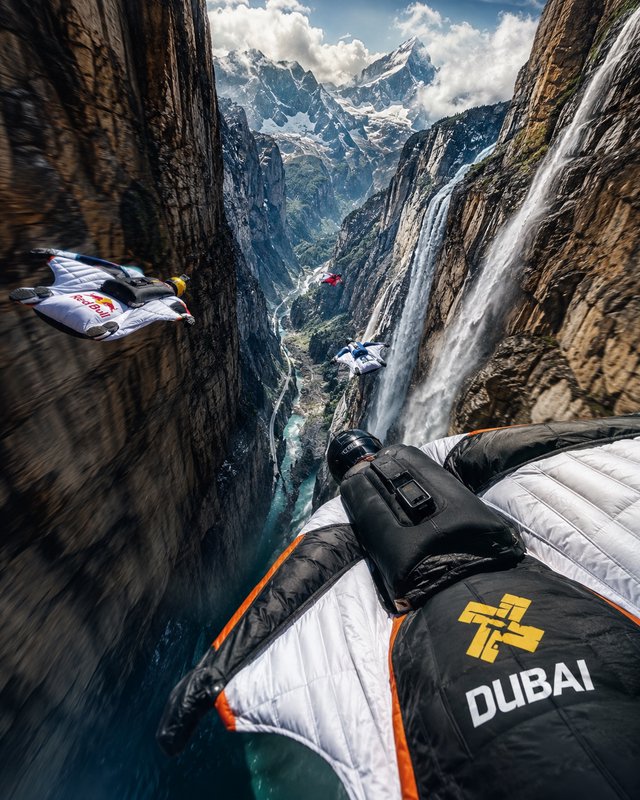POV shot of a group of wingsuit flyers gliding through a steep rocky canyon beside cascading wate...