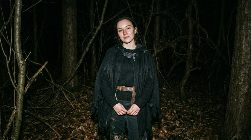 Young woman in a tattered dark cloak stands in a shadowy night forest looking toward the camera