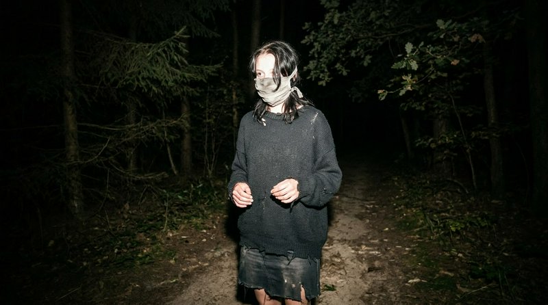 Eerie masked figure in tattered clothing stands on a dark forest path illuminated by flash at night