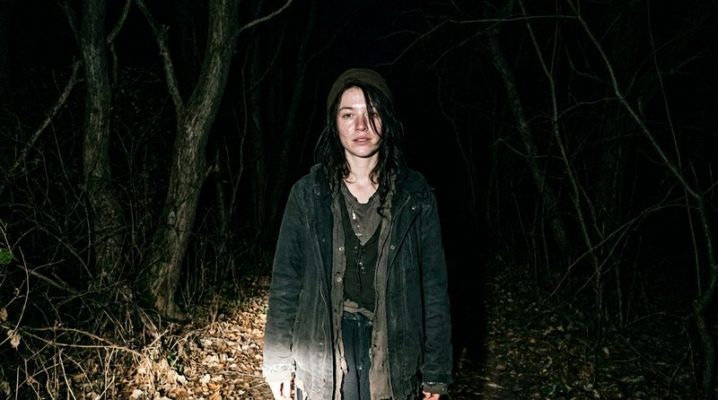 Disheveled woman in worn layered clothing stands illuminated on a dark forest path at night
