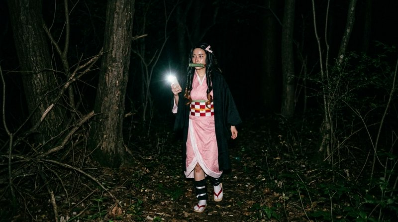 Nezuko Kamado cosplayer walks through a dark nighttime forest holding a glowing phone for light