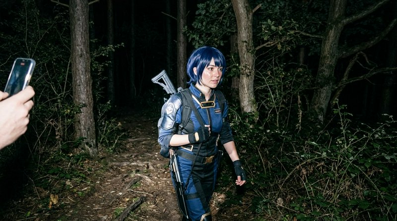 Fallout Vault Dweller cosplayer stands on a dark forest trail as a person takes a photo with a sm...